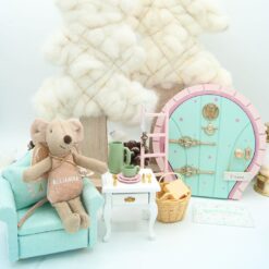 Muñeco Ratoncito Pérez Rosa personalizado con nombre de un niño sentado en sofá miniatura verde, una mesa blanca con una mini vajilla rosa y verde y cubiertos dorados. Puerta del Ratoncito Pérez rosa/verde con escalera, felpudo a juego y un cesto con queso al lado.